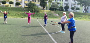 Grupa pięciu osób ćwiczy na boisku z kijkami. 